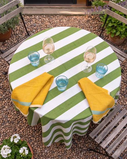 A round outdoor dining table covered with a green and white striped tablecloth, set with two yellow napkins, blue drinking glasses, and wine glasses on a pebble-covered patio.