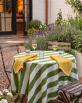 A round outdoor dining table set in a garden patio scene, covered with a green and white striped tablecloth and styled with two yellow napkins, two wine glasses, and two clear drinking glasses on wooden folding chairs.